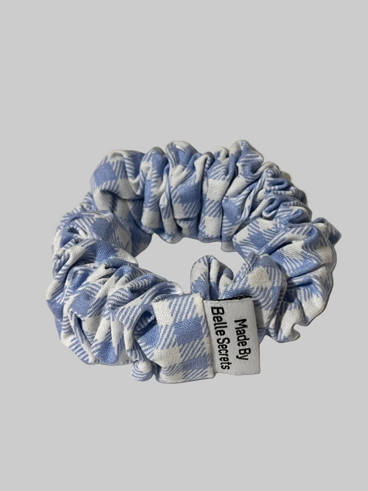 A fabric hair scrunchie with a light blue and white gingham pattern, featuring a white paper label that reads 'Made By Belle Secrets'.