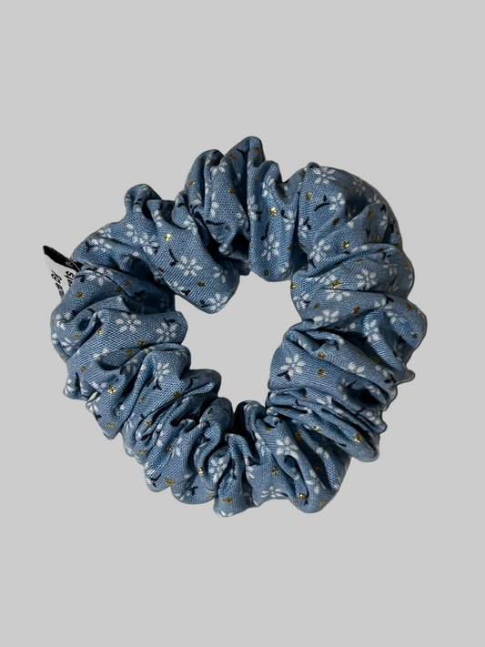 A fabric scrunchie in a dusty blue color with a white floral pattern and small blue accents, resting on a light blue textured fabric background.